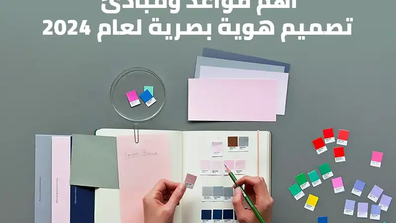 يد مصمم تمسك بقلم وتقوم بعمل تصميم على كتاب على خلفية رمادية مكتوب عليها أهم قواعد ومبادئ تصميم هوية بصرية لعام 2024